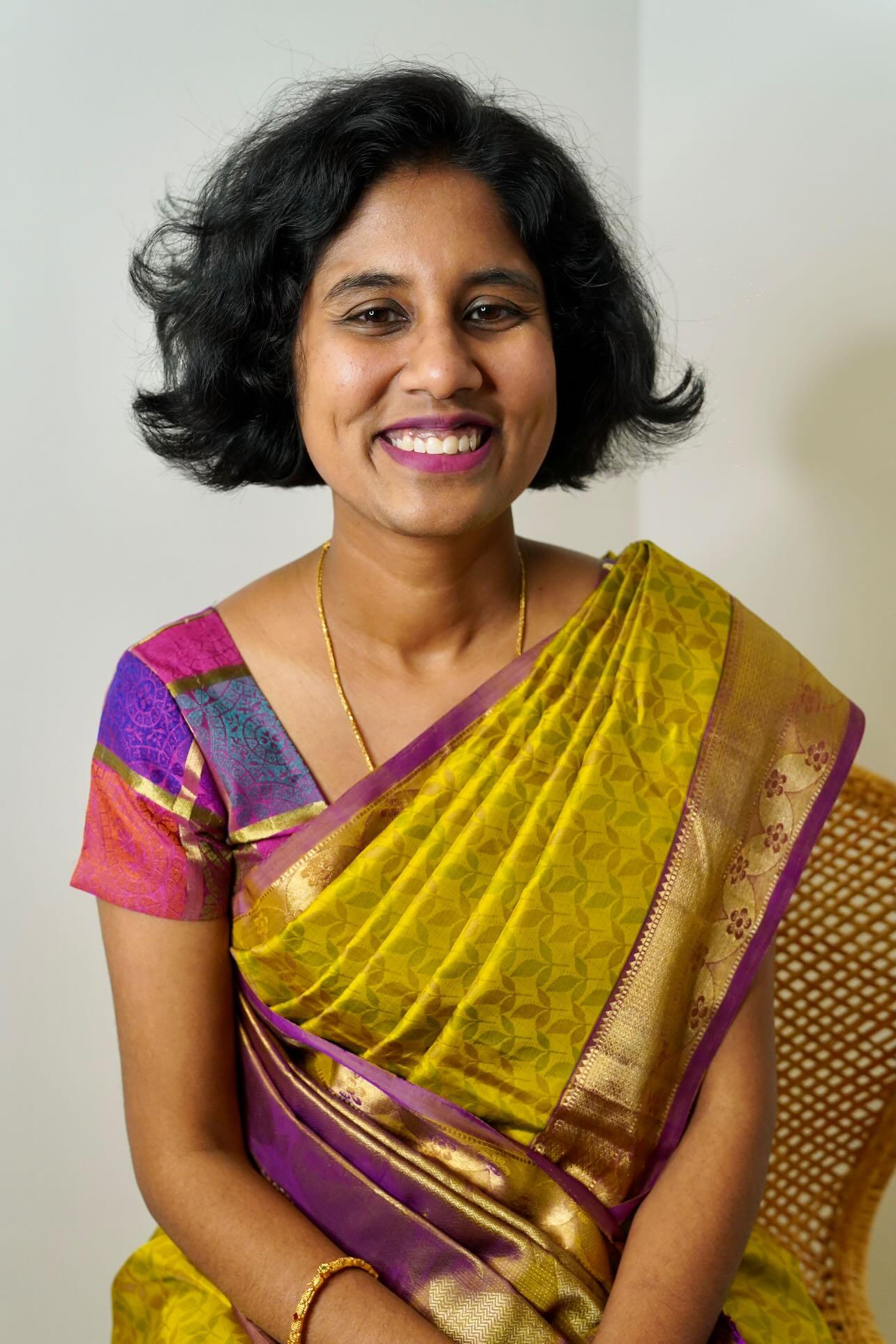 Person wearing a green silk saree with a purple border, featuring short hair and purple lipstick.